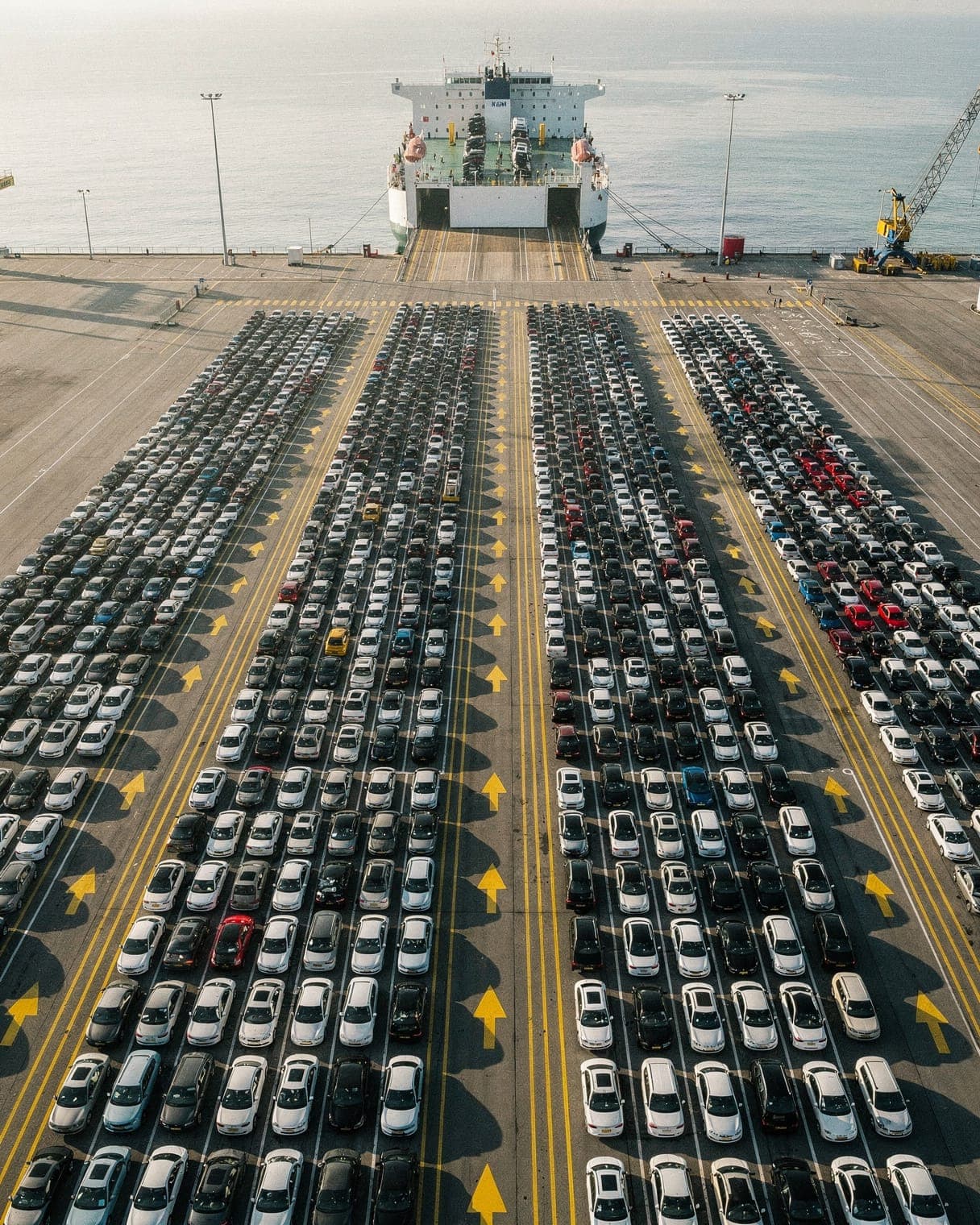 Rows of newly arrived cars at a UK port, lined up beside a car carrier ship